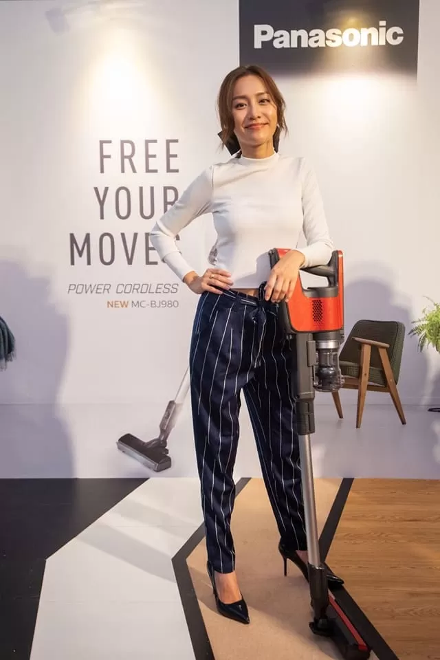 a woman standing with a bright red Panasonic cordless vacuum cleaner used as a support in front of a backdrop on the left. Another woman with a white Panasonic cordless vacuum cleaner is sitting on a flight of wooden stairs.